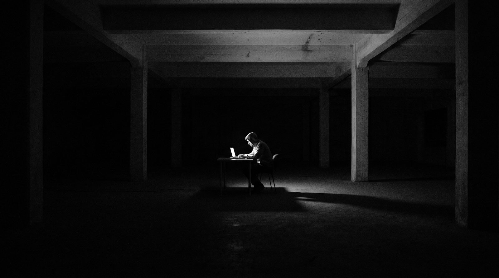 A lone figure working at a desk lit only by a laptop, in a vast dark empty space