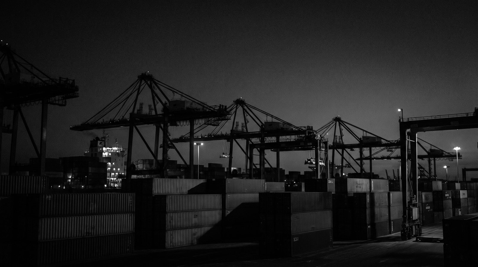 A cargo shipping port at night in silhouette, cranes and containers against a dark sky